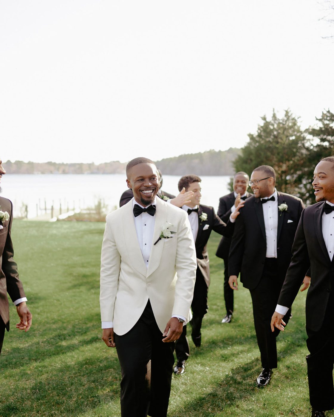 Groom in a white dinner jacket walking the lakeside lawn with his groomsmen